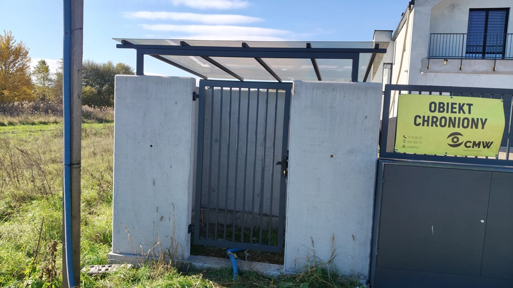 Wiata na śmieci z betonowymi słupami i metalową furtką pod zadaszeniem